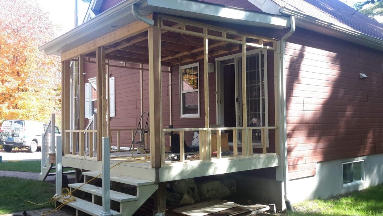 Un porche latéral d'habitation en construction, avec une structure en bois, un escalier et des poutres apparentes, sur une maison en briques.