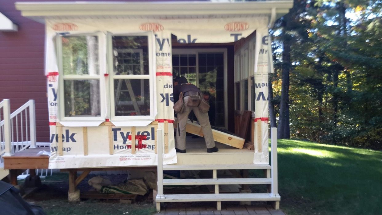 Une personne en tenue de travail transporte une grosse poutre en bois dans une extension de maison inachevée, recouverte d'une bâche Tyvek.