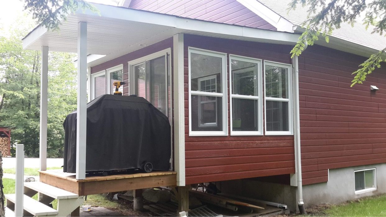 Une maison rouge aux boiseries blanches, un porche couvert avec un barbecue et une rangée de fenêtres au-dessus d'une terrasse inachevée.