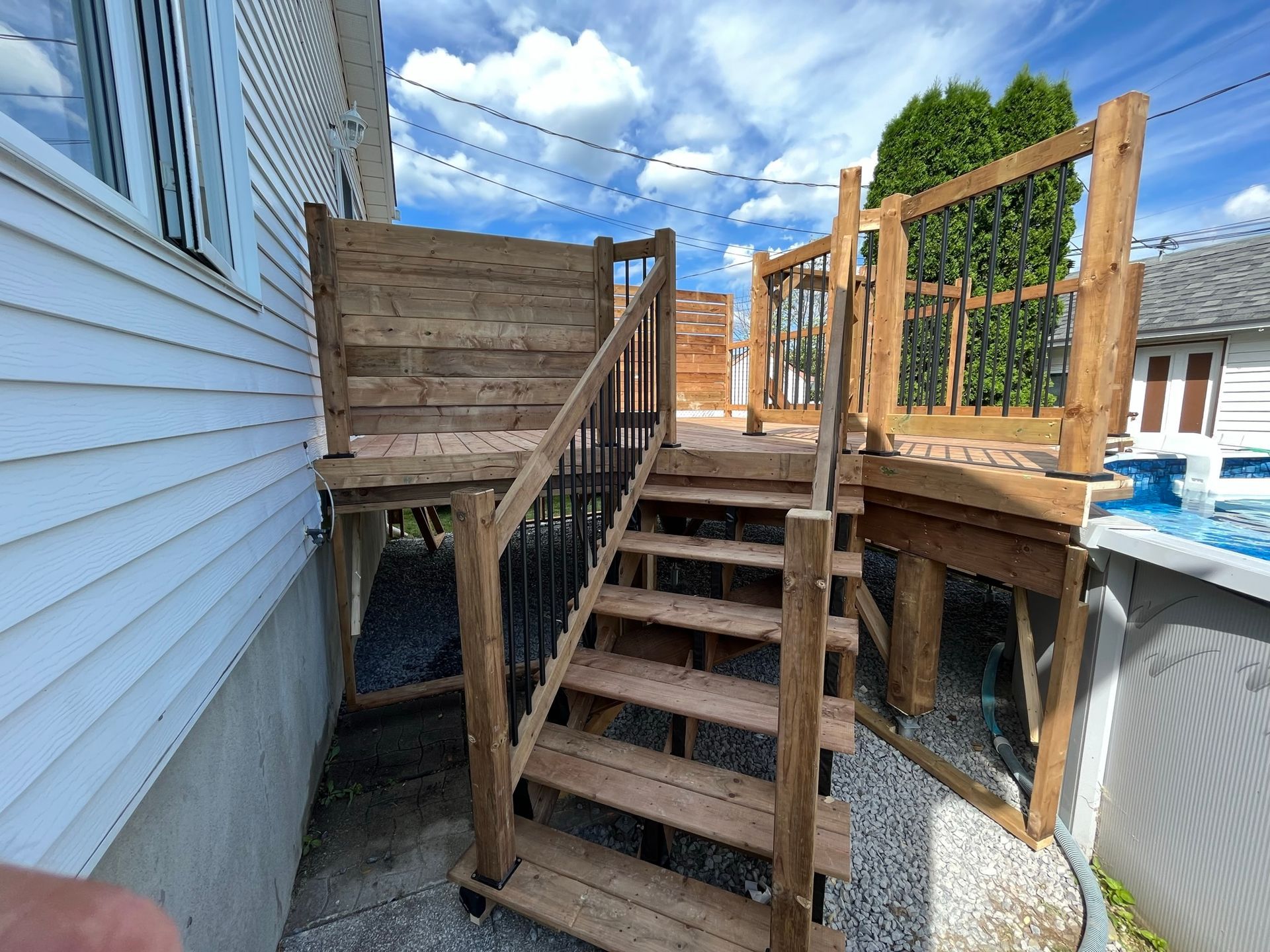 Un escalier en bois mène à une terrasse en bois jouxtant une maison, avec une rambarde en métal et une piscine attenante.