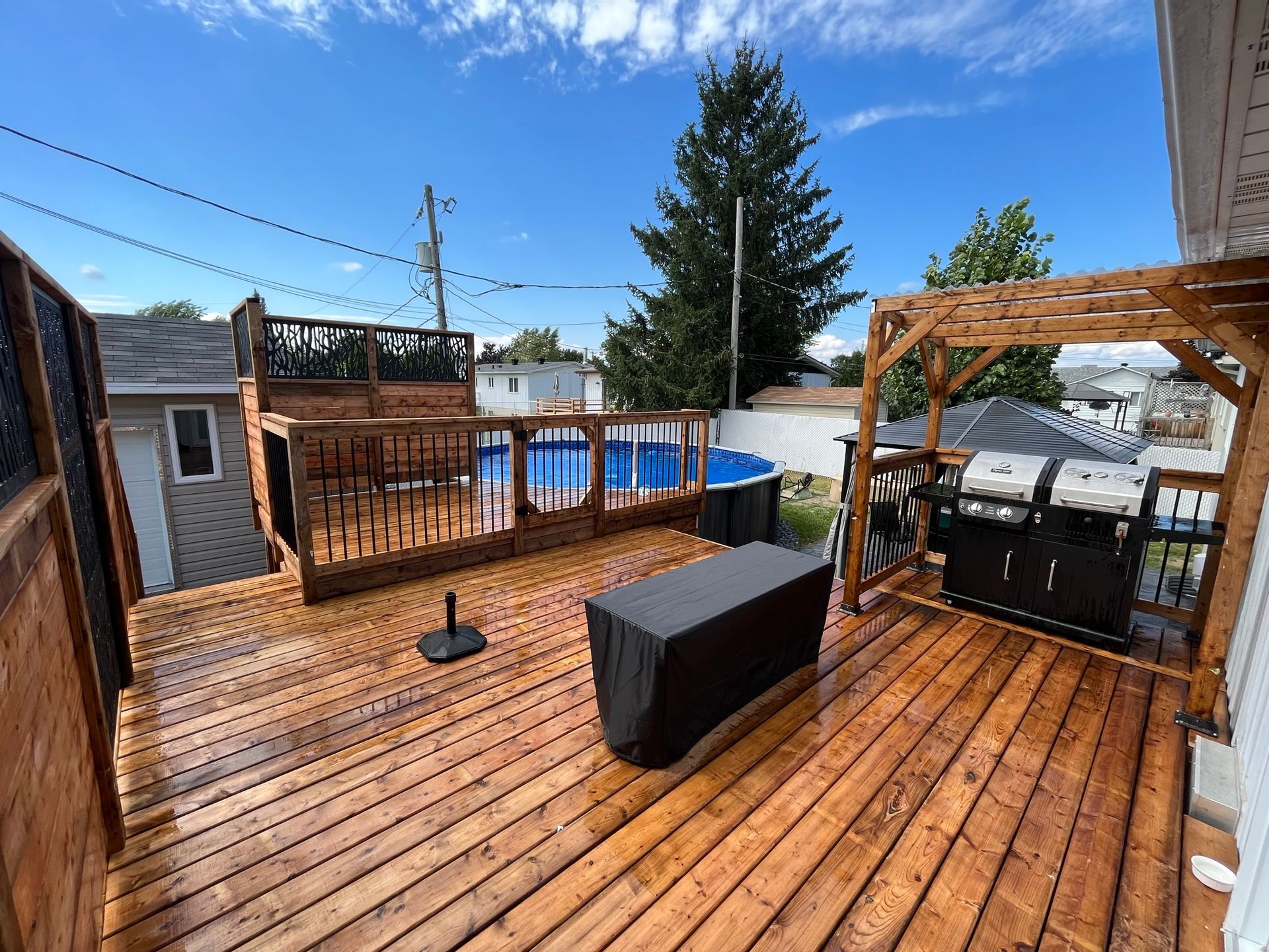 Une terrasse en bois avec piscine hors-sol bleue, pergola en bois, grill noir, mobilier de jardin et cl&ocirc;ture, sous un ciel bleu