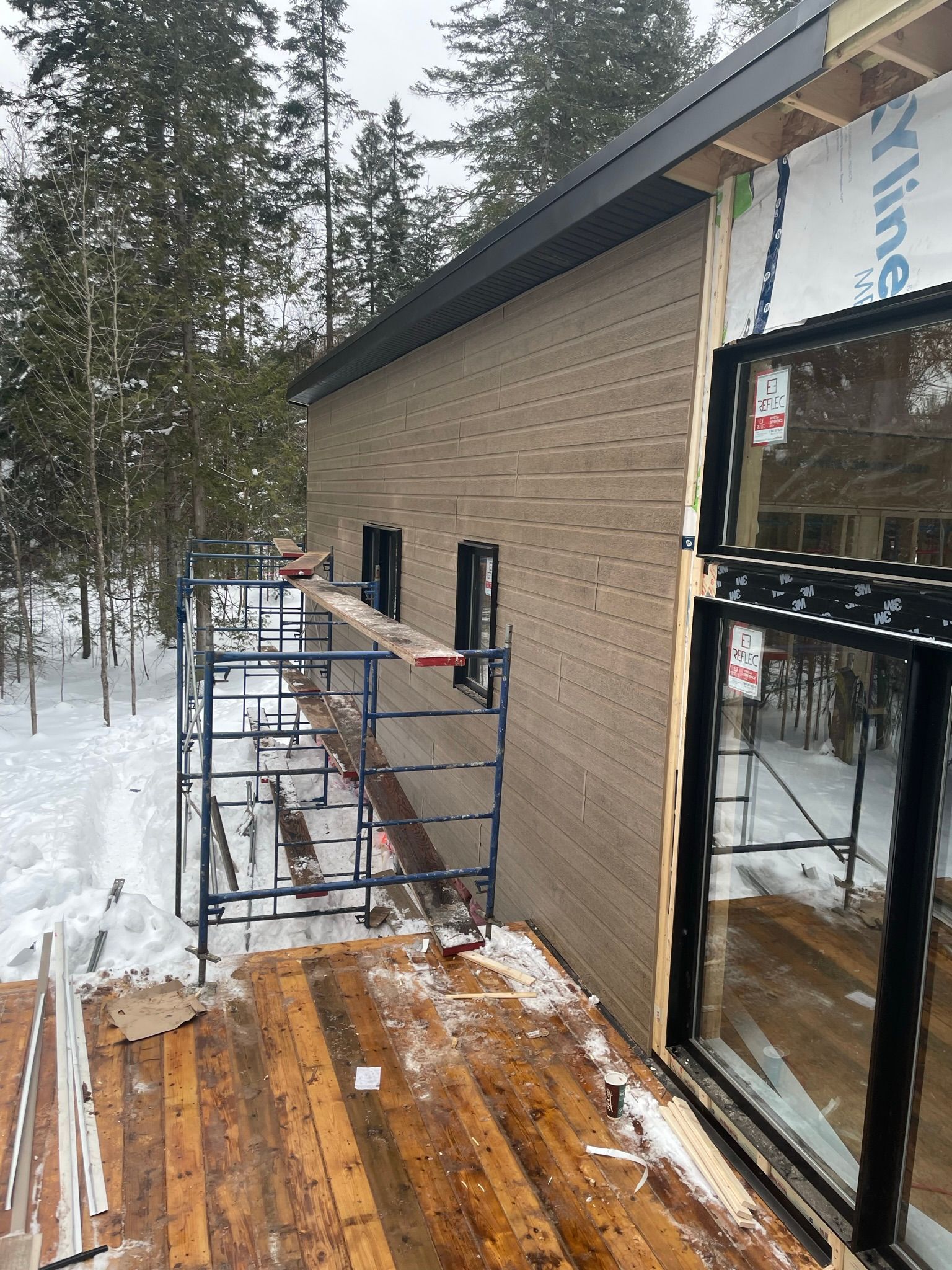 Une maison est en construction dans la neige avec une terrasse en bois devant.