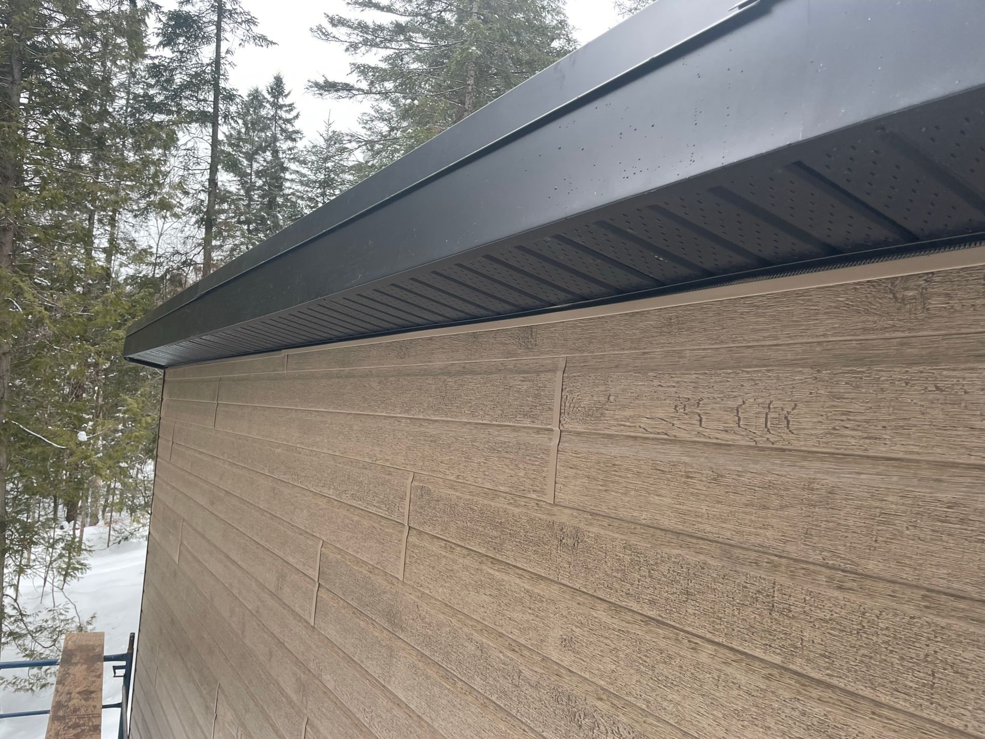 Mur de garage en bois clair avec toit en m&eacute;tal gris, entour&eacute; de sapins enneig&eacute;s.