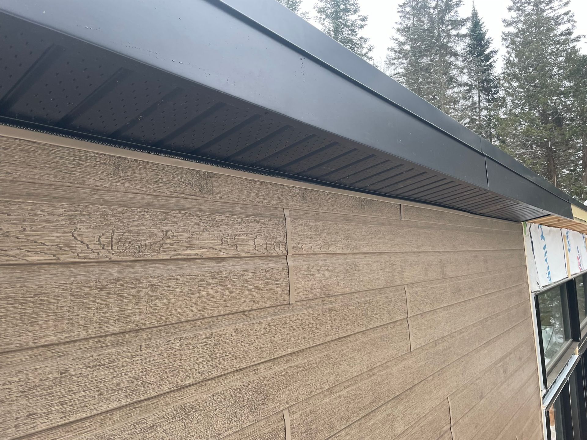 Mur en bois horizontal avec surplomb m&eacute;tallique gris, devant une for&ecirc;t de conif&egrave;res.