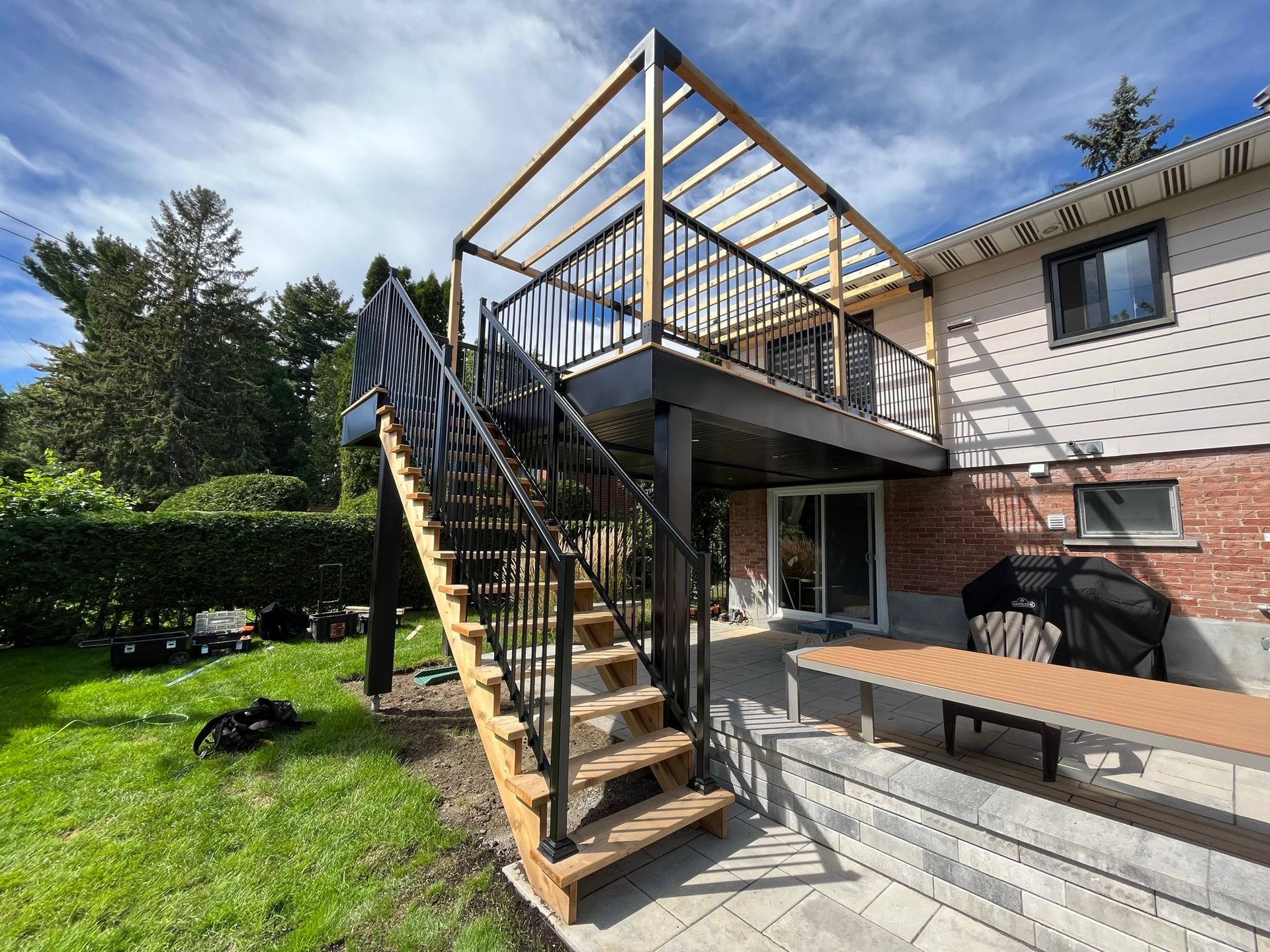 Une terrasse surélevée en bois avec des balustrades noires et une structure de pergola en bois, reliée par des escaliers à un patio donnant sur le jardin.