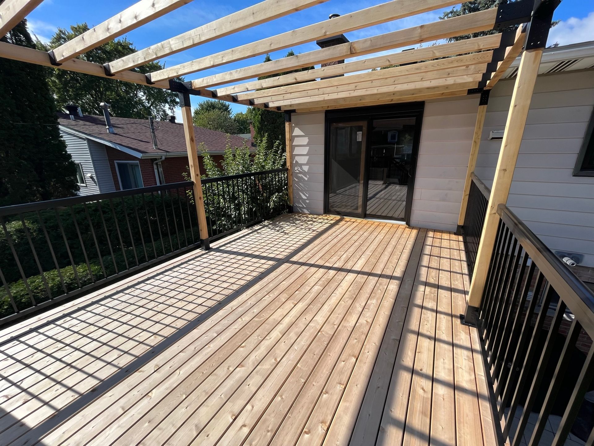 Terrasse en bois avec pergola ajour&eacute;e et garde-corps noir, adjacente &agrave; une maison.