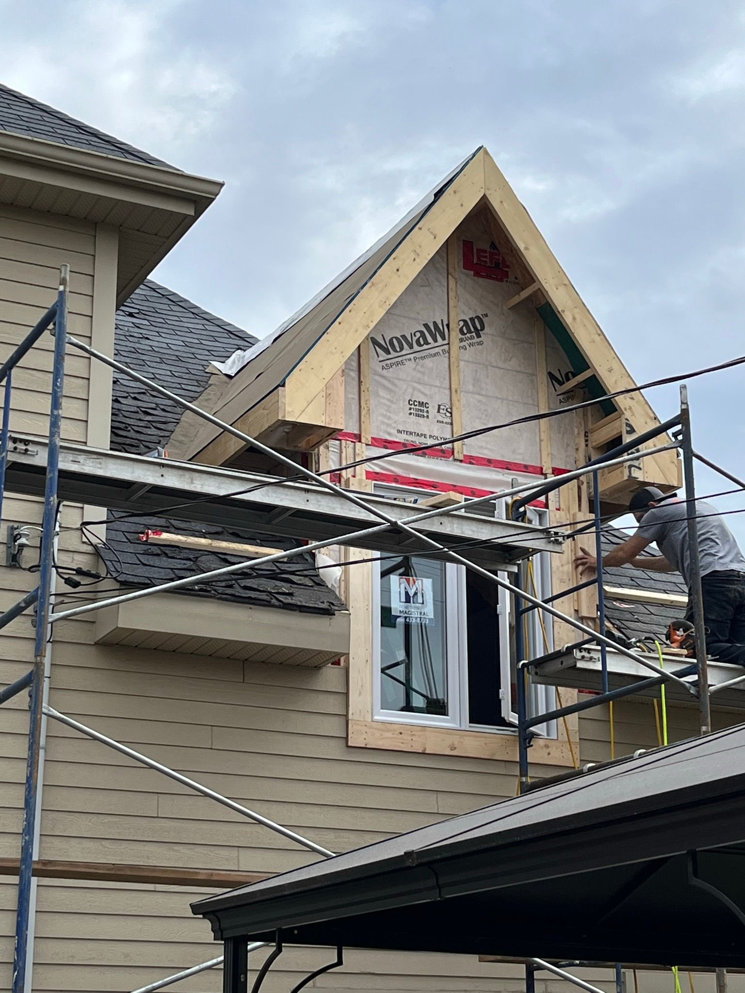 Un ouvrier, perché sur un échafaudage, installe des moulures en bois autour d'une fenêtre à pignon au deuxième étage d'une maison en construction.