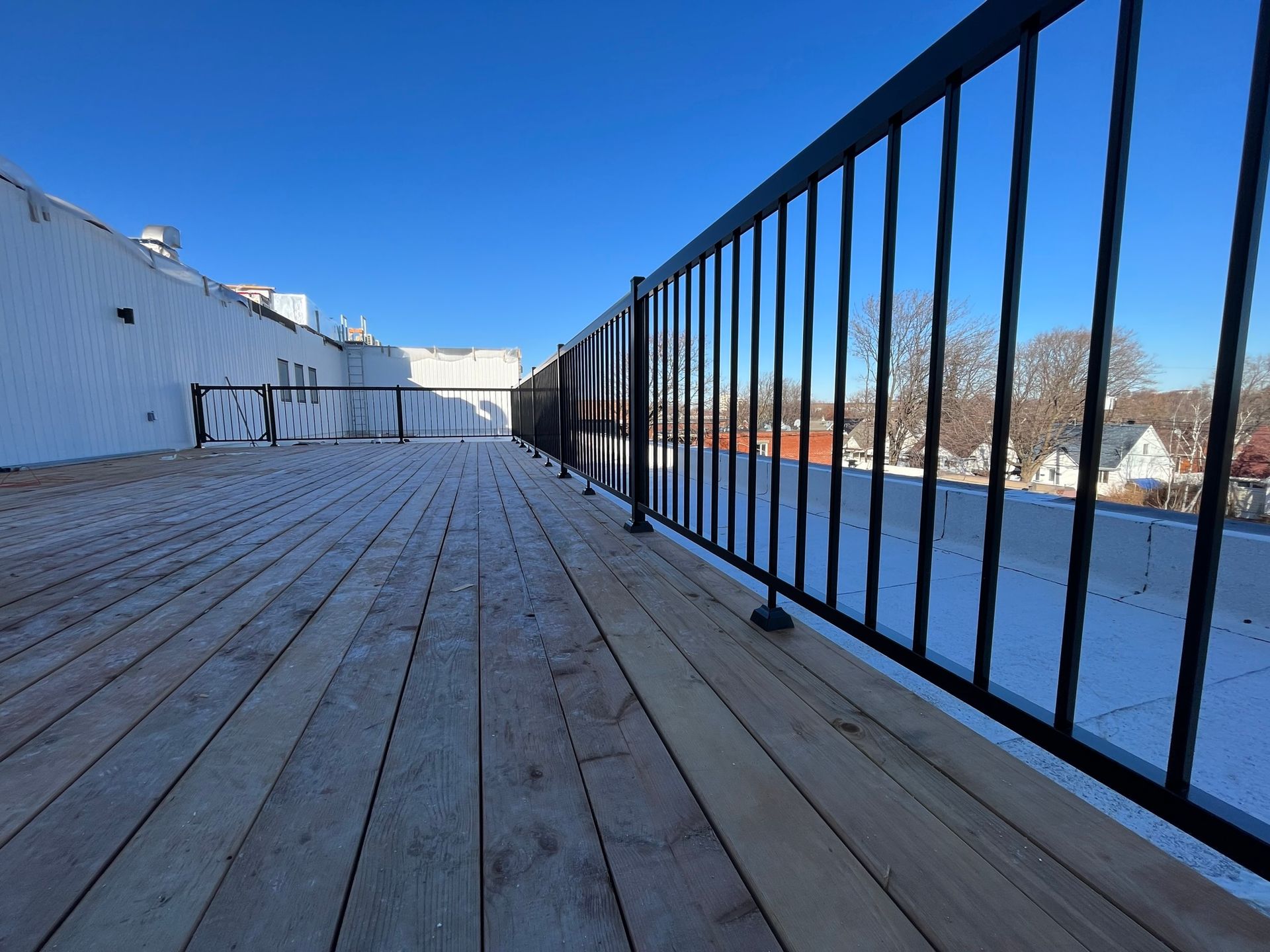Une terrasse en bois sur le toit, dotée d'une rambarde en métal noir, sous un ciel bleu azur.