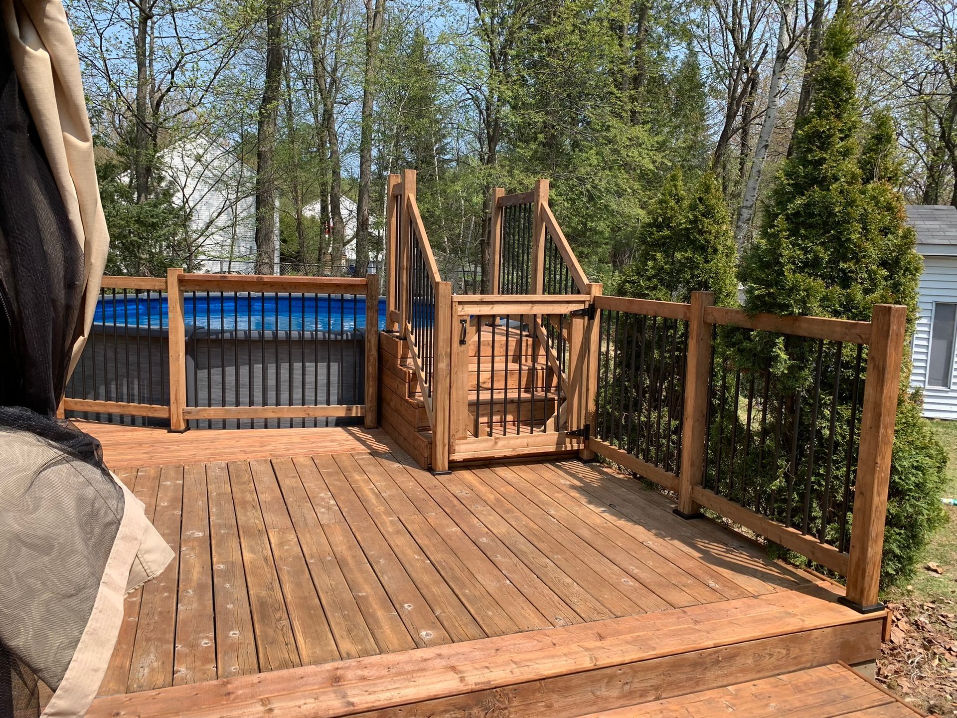 Une terrasse en bois avec un portillon de sécurité menant à une piscine hors sol, entourée d'arbres et d'un auvent beige au premier plan.