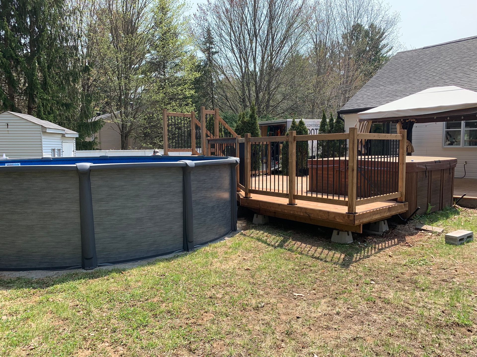 Une piscine hors sol ronde, aux panneaux gris, jouxte une terrasse en bois avec un jacuzzi, le tout dans un jardin ensoleillé et gazonné.