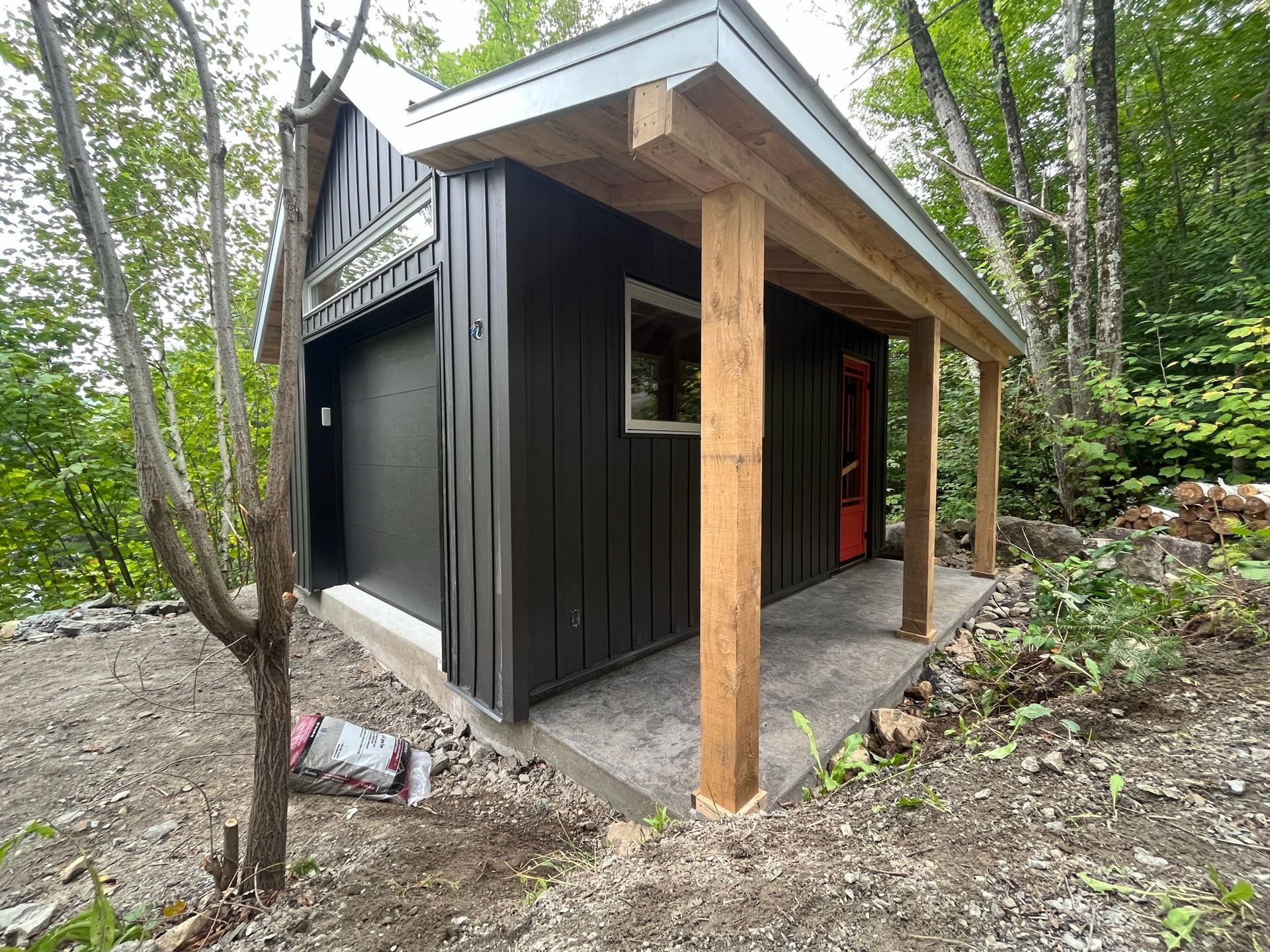 Un petit garage noir avec un porche en bois et une porte rouge.