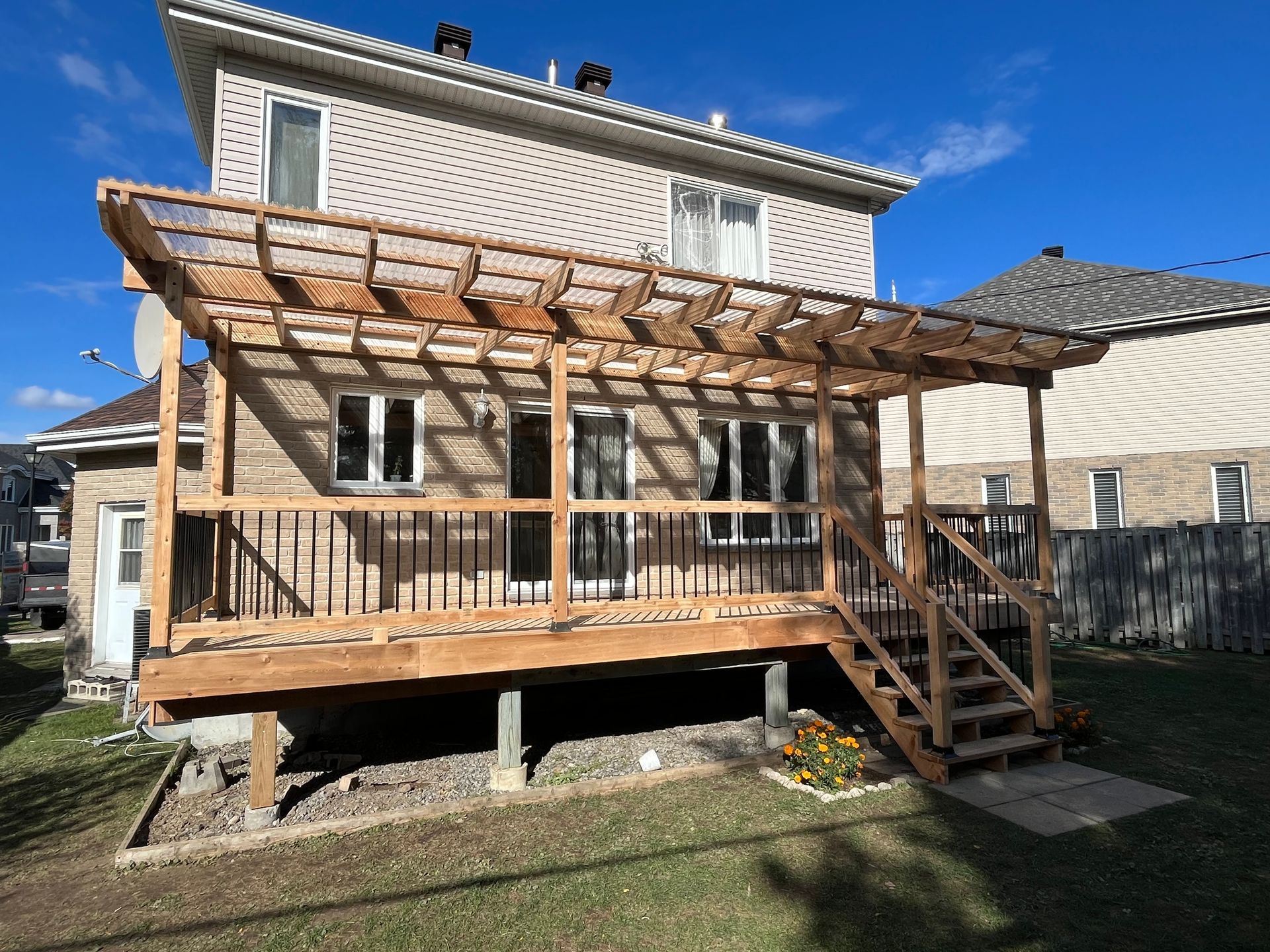 Une maison en briques beiges avec une terrasse en bois et une pergola nouvellement construites dans le jardin, sous un ciel bleu.