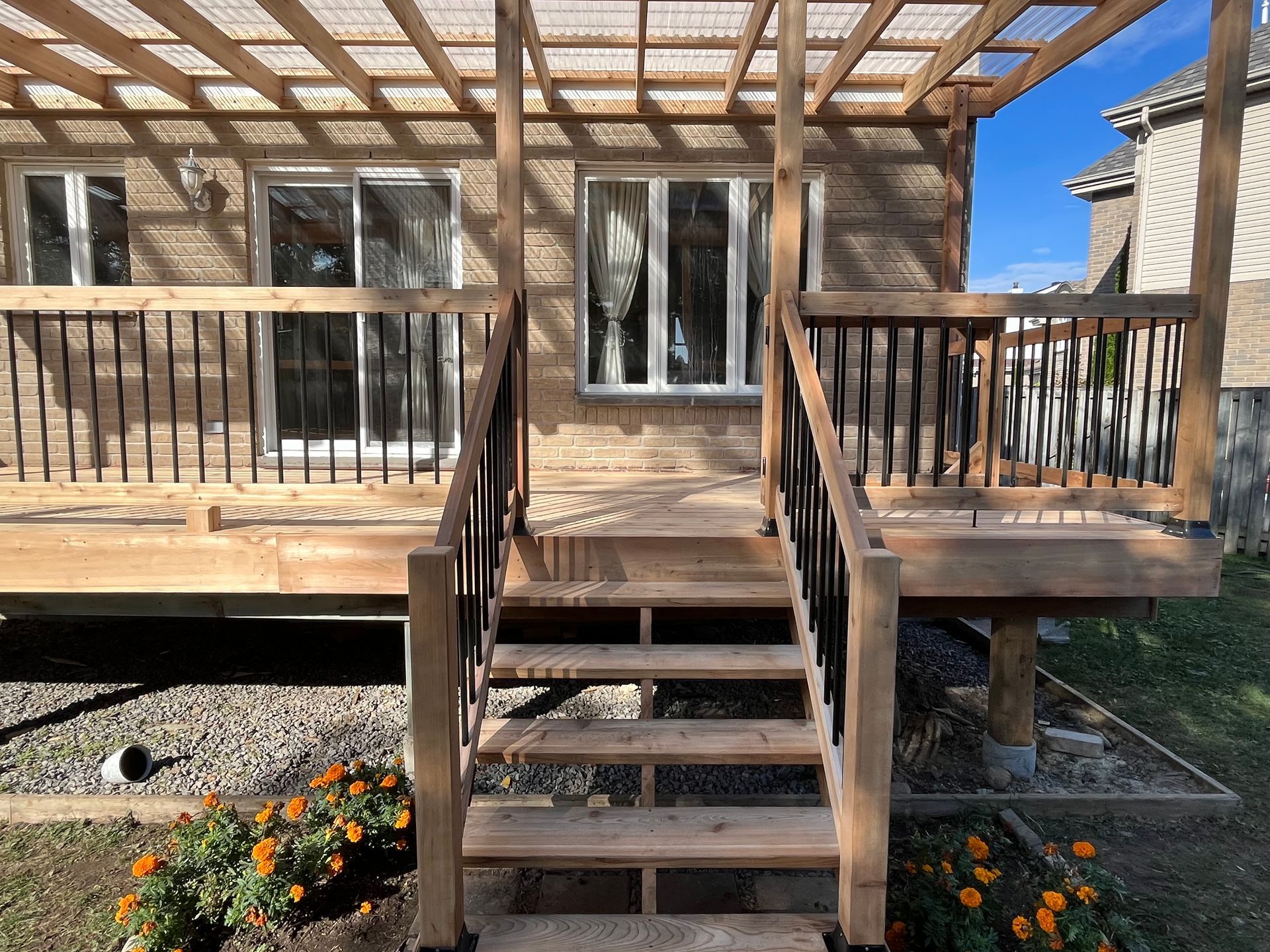 Une terrasse en bois avec escalier, rambarde noire et pergola, adossée à l'arrière d'une maison à deux fenêtres.