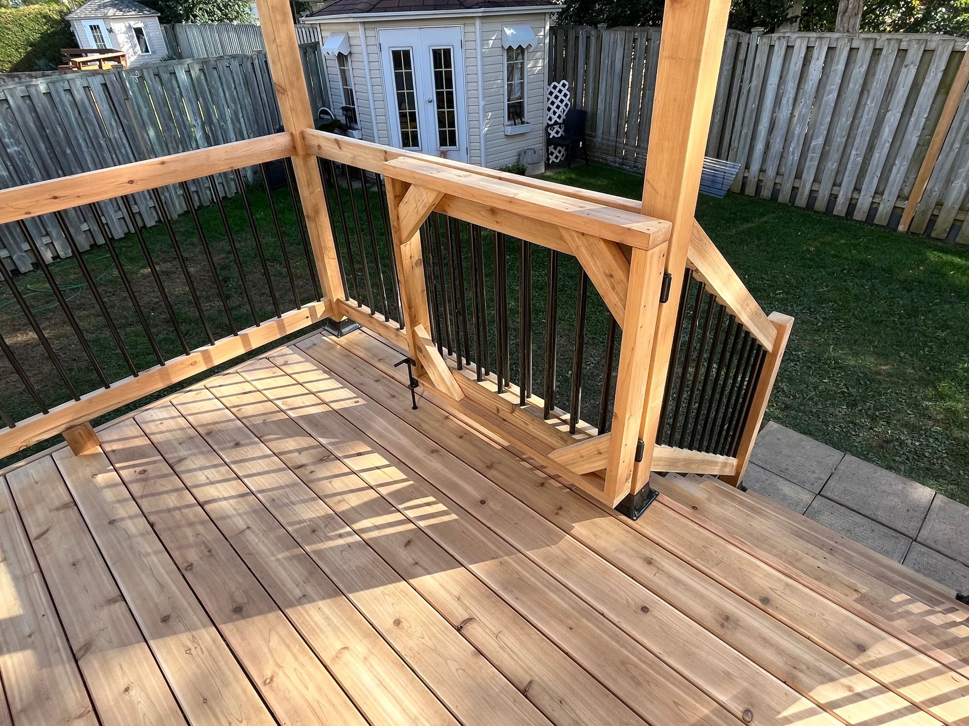 Une nouvelle terrasse en bois avec rambarde, balustres en métal noir et portail, donnant sur un jardin avec un abri de jardin et une clôture.