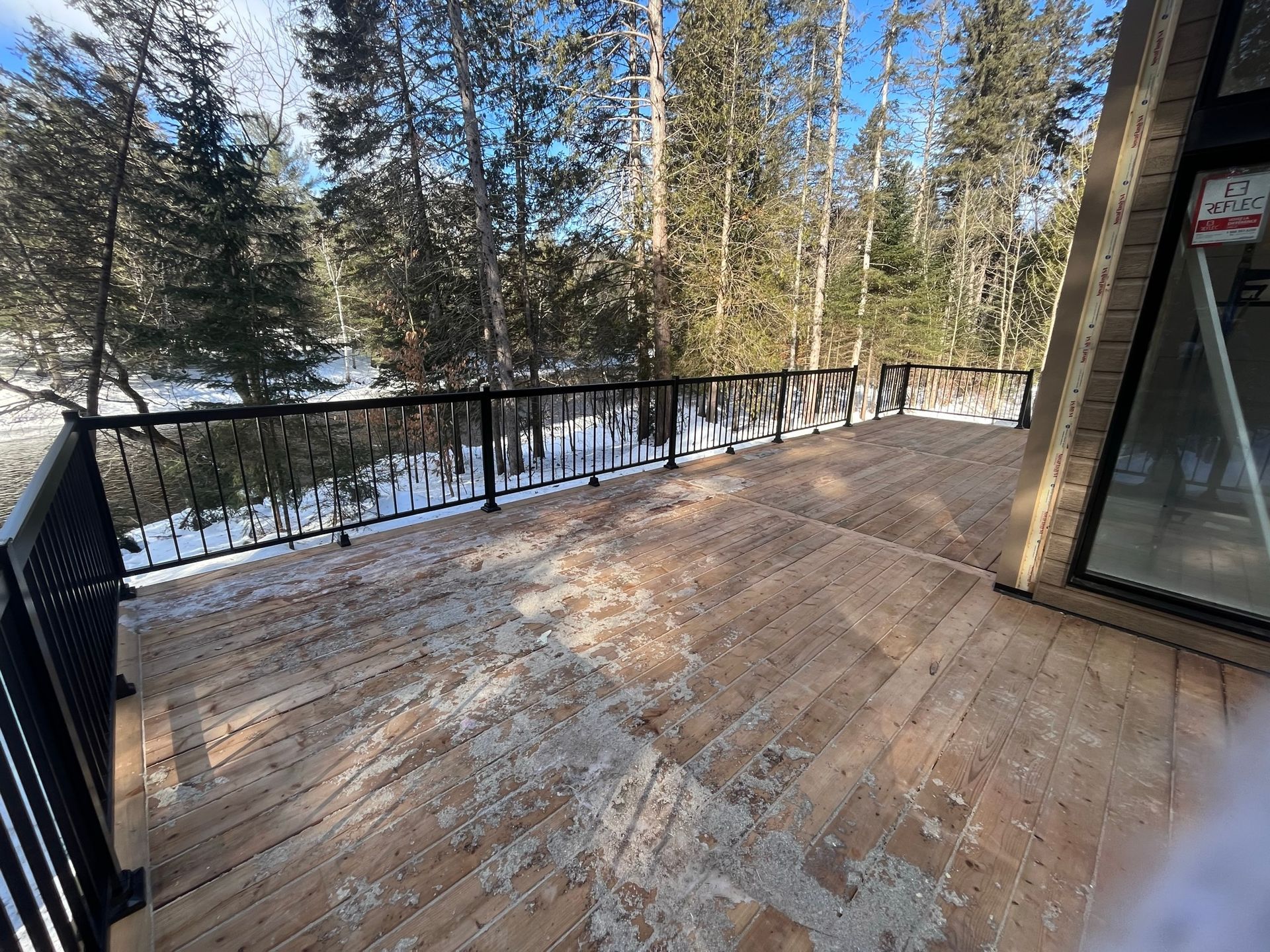 Une terrasse en bois avec une rambarde noire surplombe une forêt enneigée par une journée d'hiver ensoleillée.