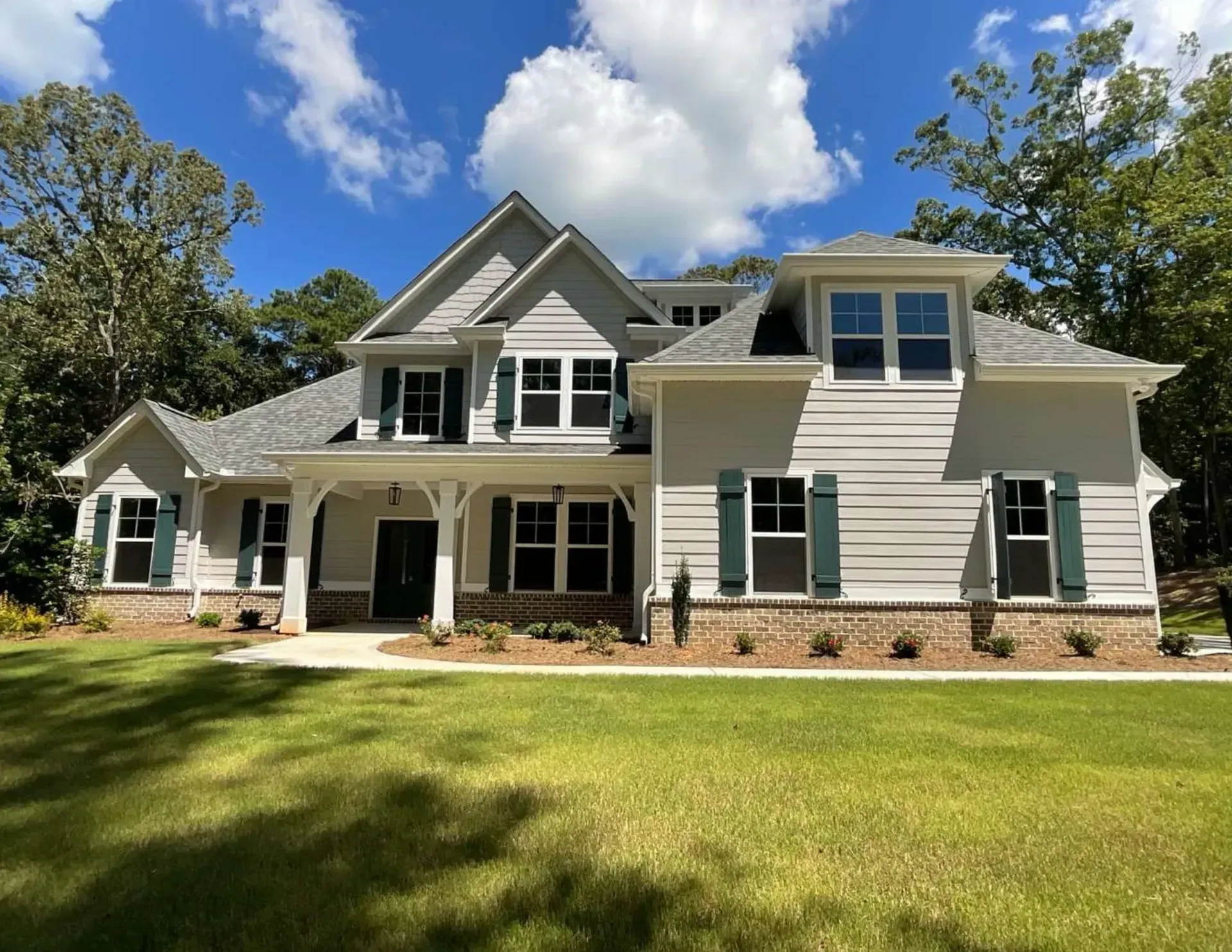Large white suburban house with green shutters, brick accents, and a wide front lawn under a blue sky