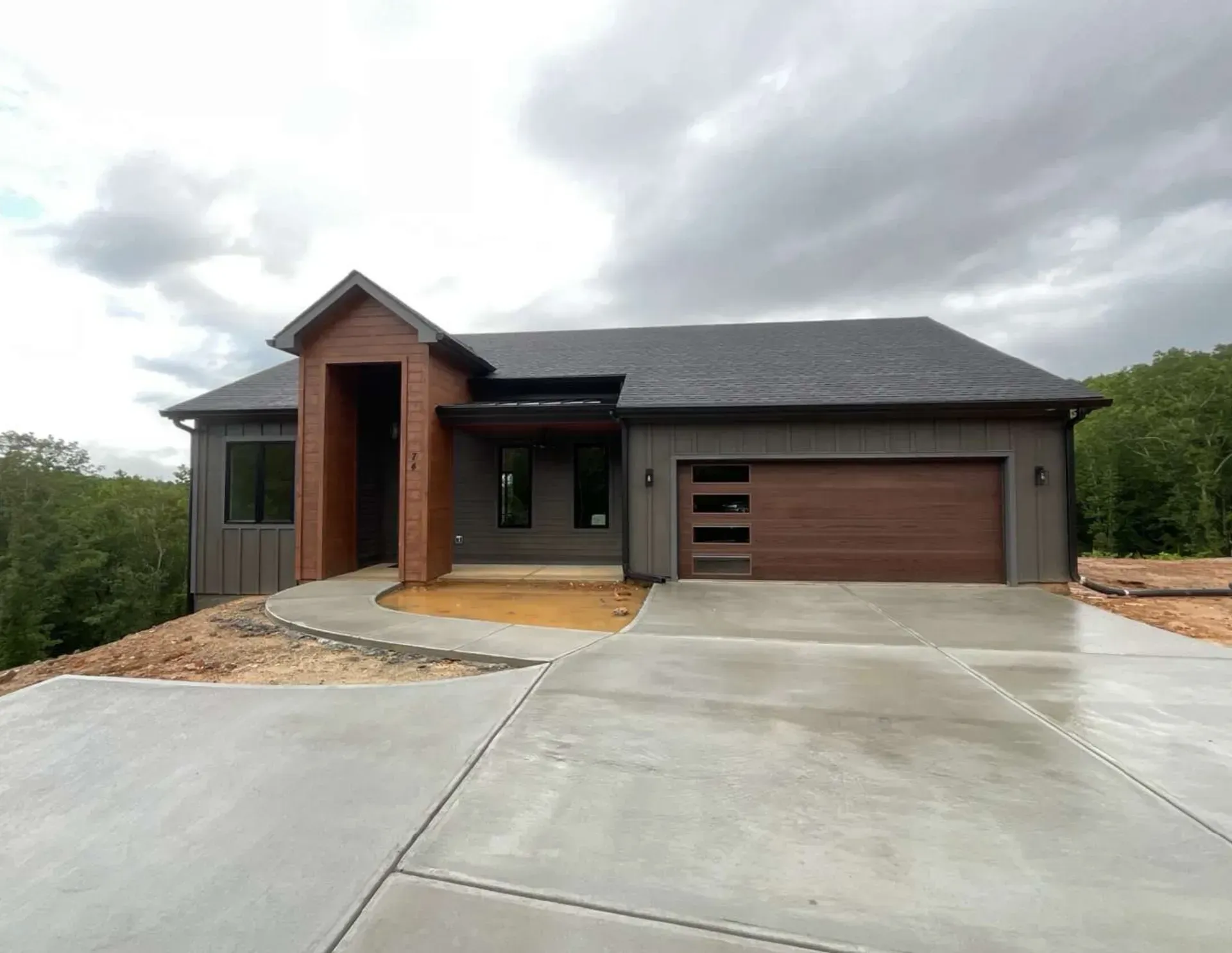 Modern single-story house with brick and wood accents, large driveway, and attached garage under cloudy sky