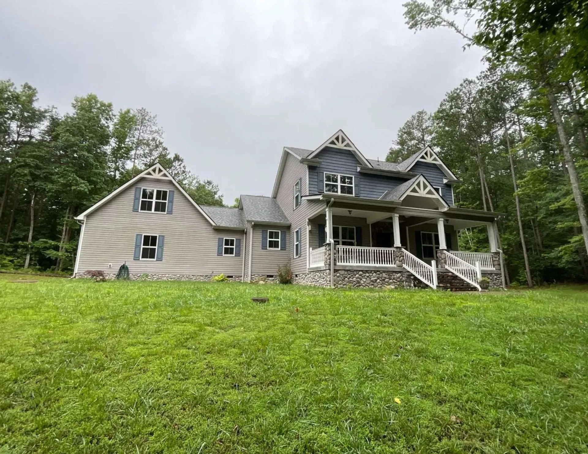 Large gray house with a covered porch on a grassy hillside, surrounded by trees under a cloudy sky
