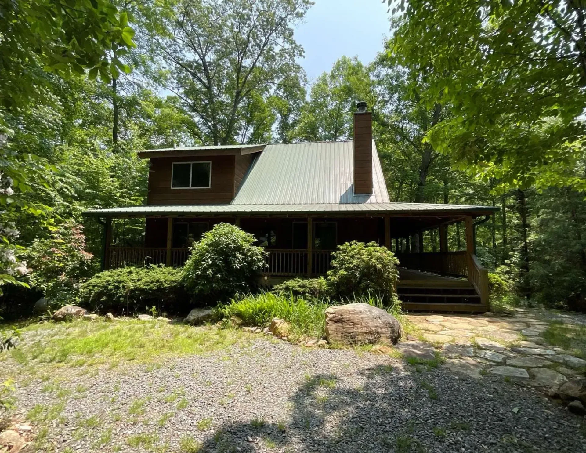 Rustic cabin with a green metal roof, front porch, and stone chimney in a wooded setting