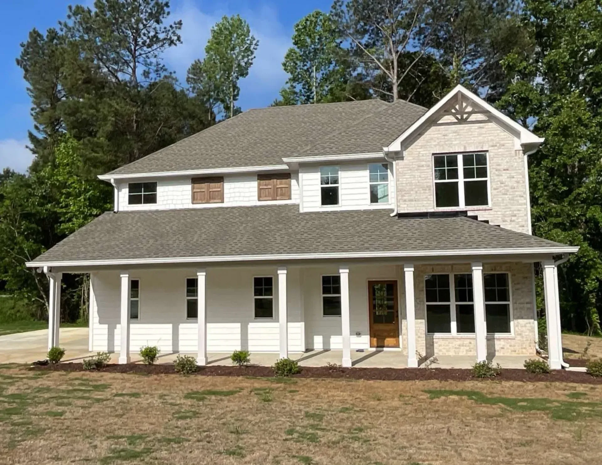 Two-story white house with a wraparound porch and gray roof, surrounded by trees on a sunny day