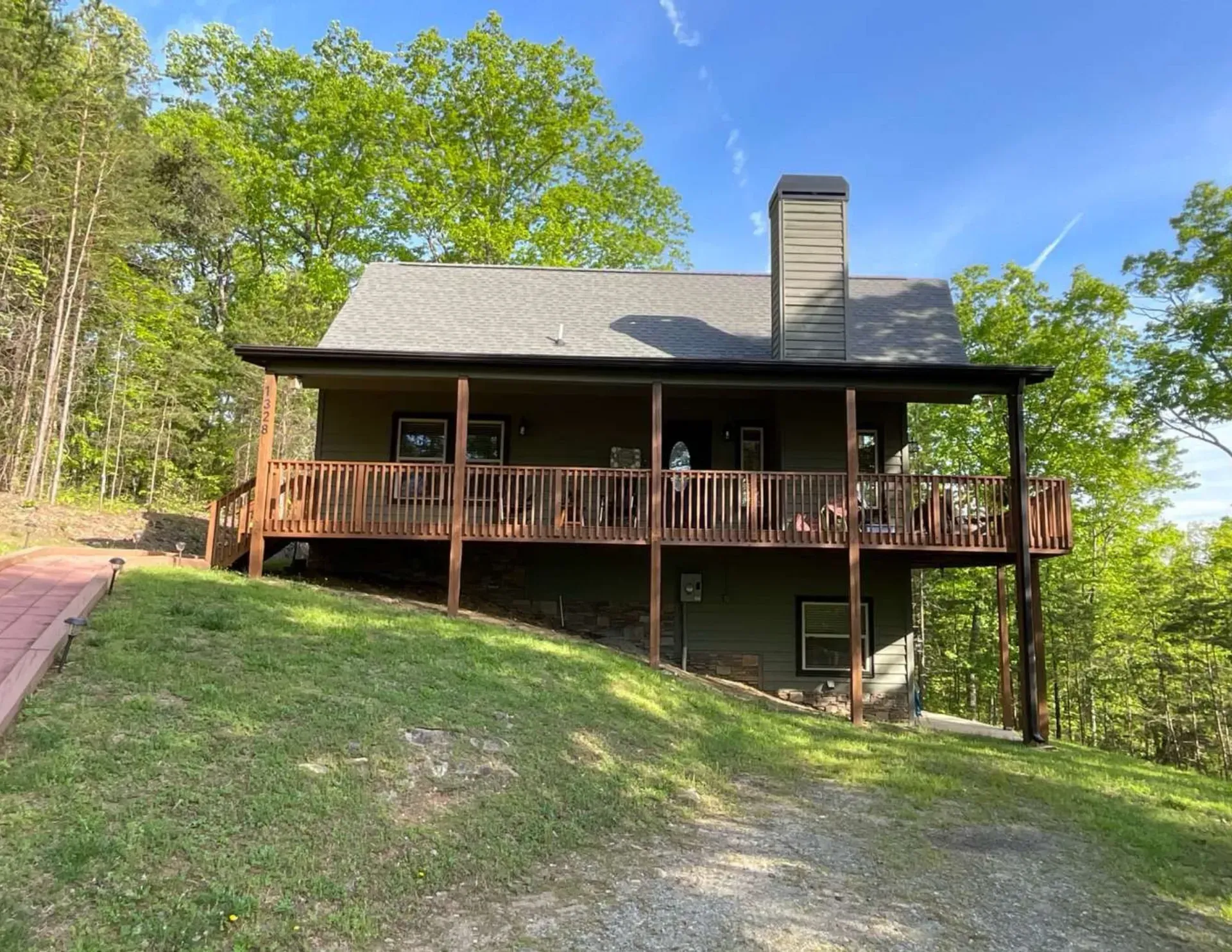 Wooden cabin with a wraparound porch on a grassy hillside, surrounded by trees under a blue sky
