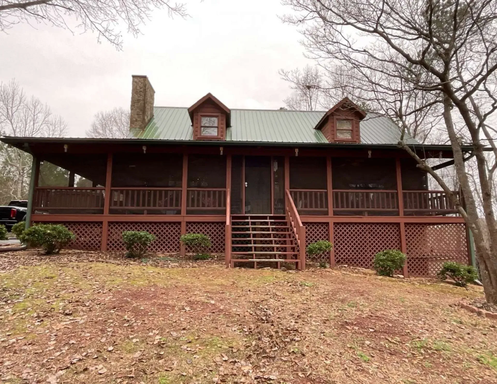 Rustic wooden cabin with a covered porch, dormer windows, and a green metal roof on a leaf-strewn lot