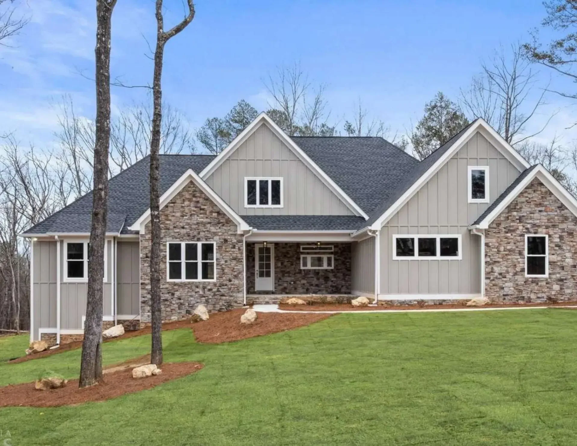 Large modern farmhouse with stone and gray siding, dark roof, and manicured green lawn under a blue sky