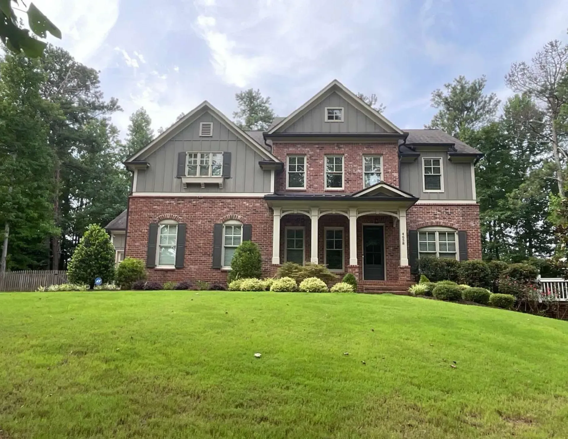 Large two-story brick house with gray siding, front porch, and a wide green lawn under cloudy skies