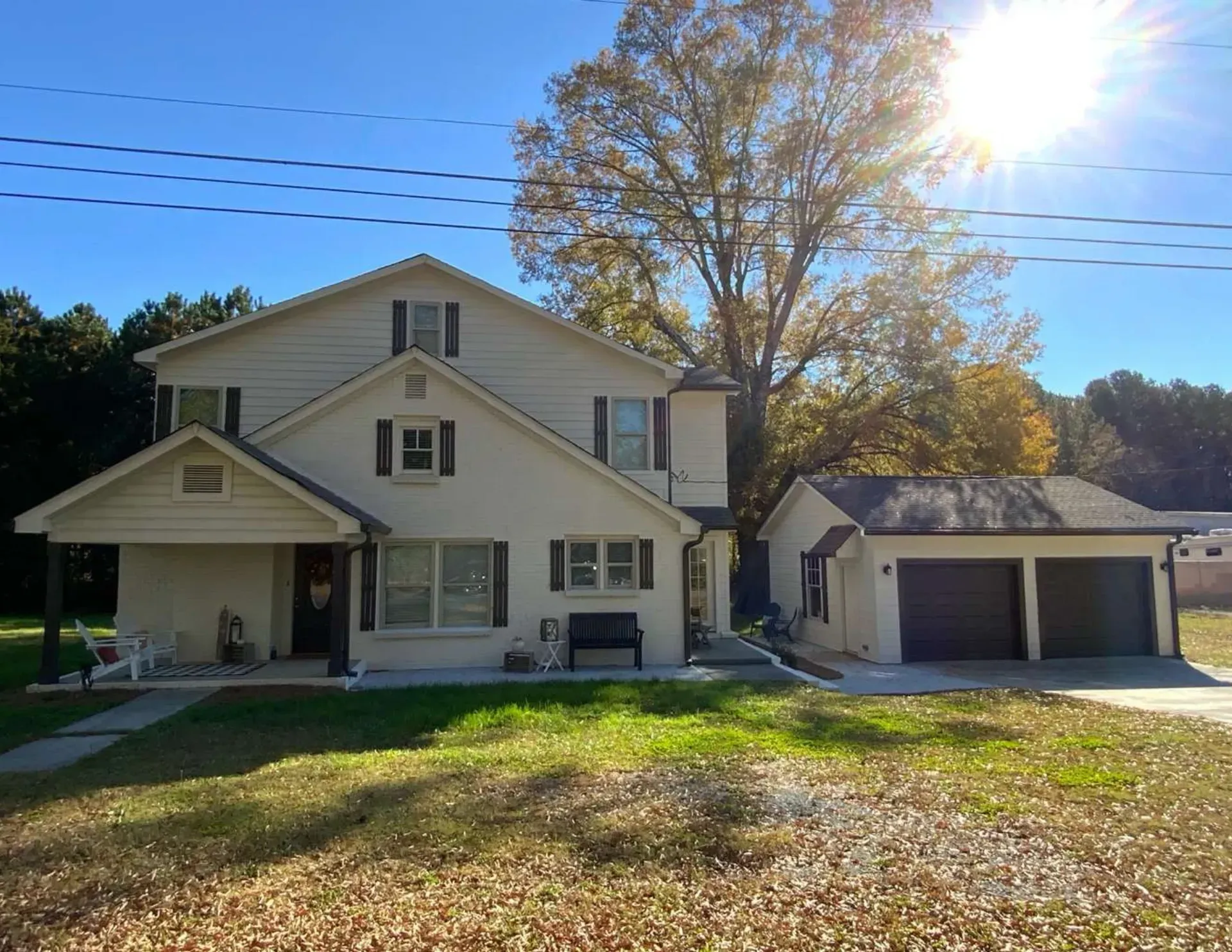White farmhouse with detached garage in autumn sunlight, front lawn covered with fallen leaves