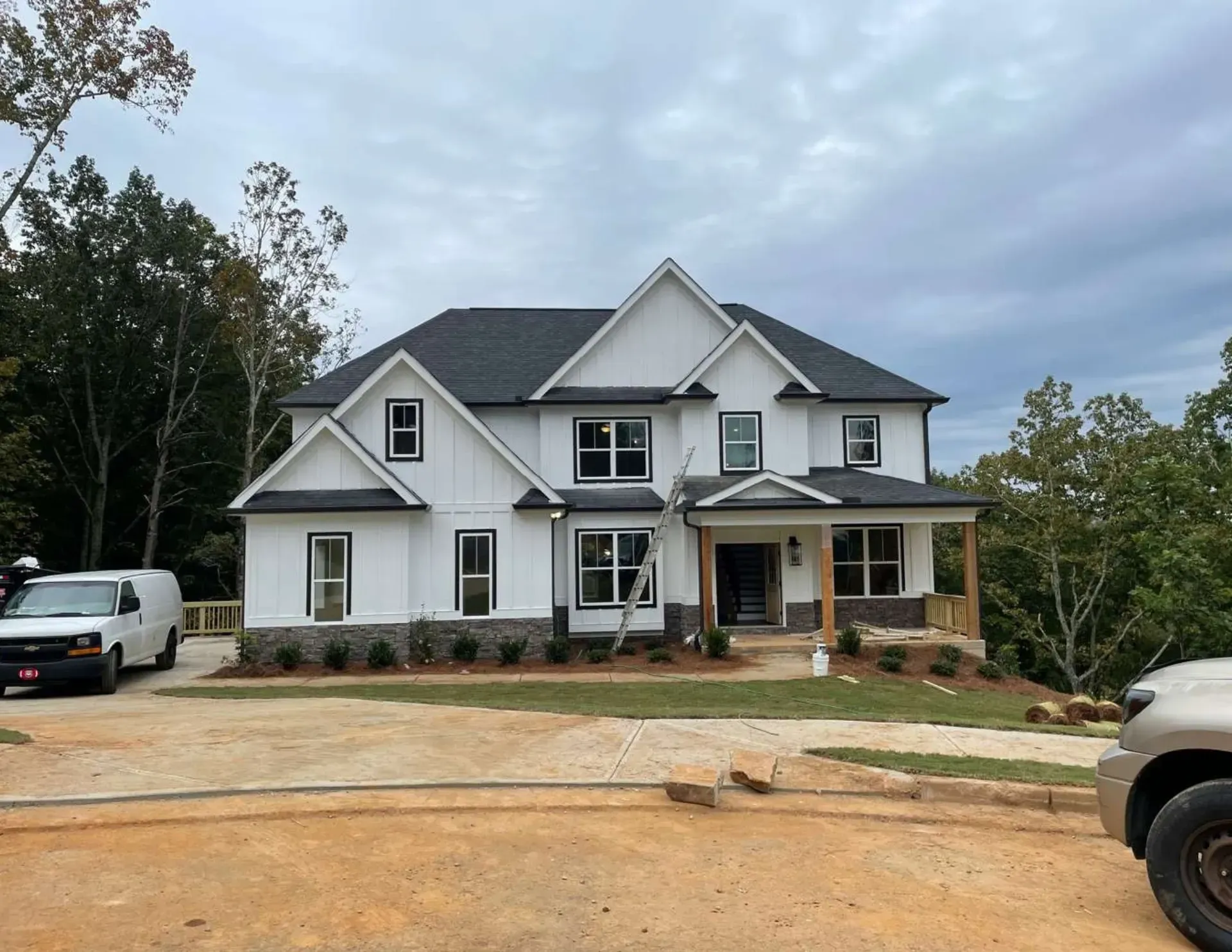 Large gray two-story house under construction with a front porch and parked vehicles on a dirt lot