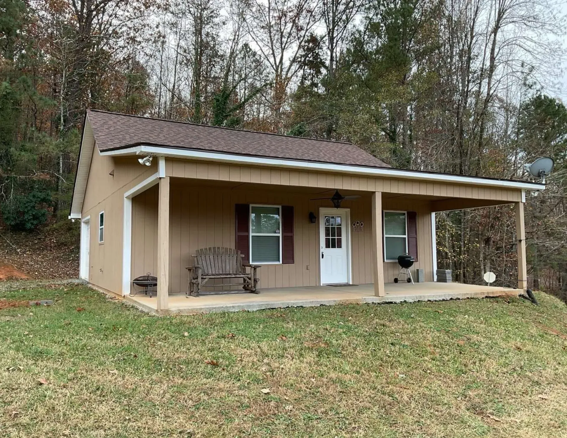 Small tan cabin with a porch and white door on a grassy lot, surrounded by trees.