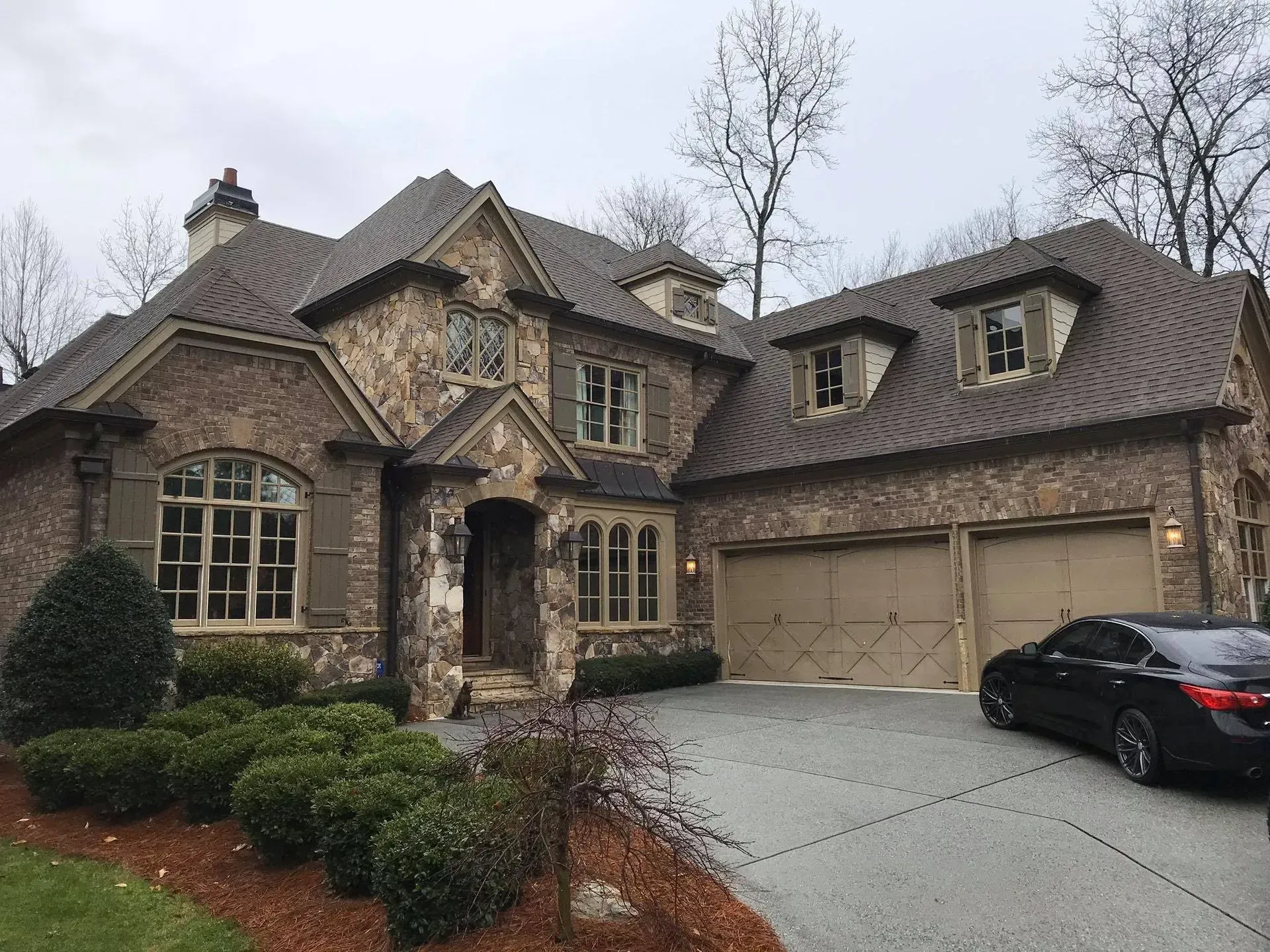 Large stone suburban house with arched entrance, attached garage, and a black car in the driveway.