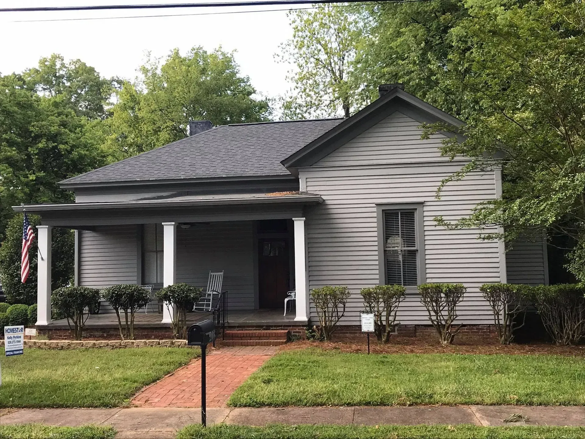 Small gray house with a covered porch, dark roof, and front lawn bordered by a sidewalk.