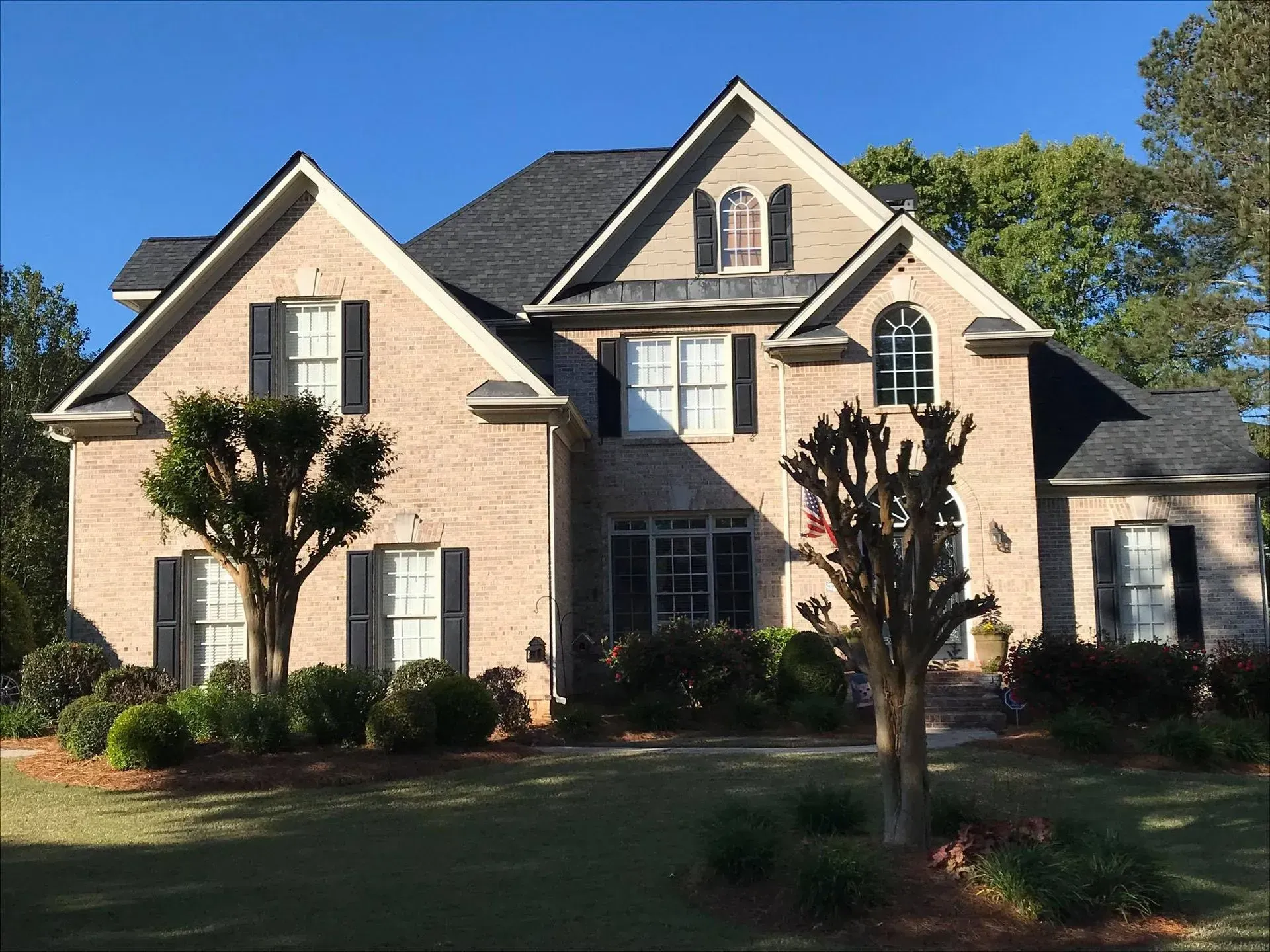Large brick suburban house with a dark roof, front lawn, and trimmed trees in bright sunlight