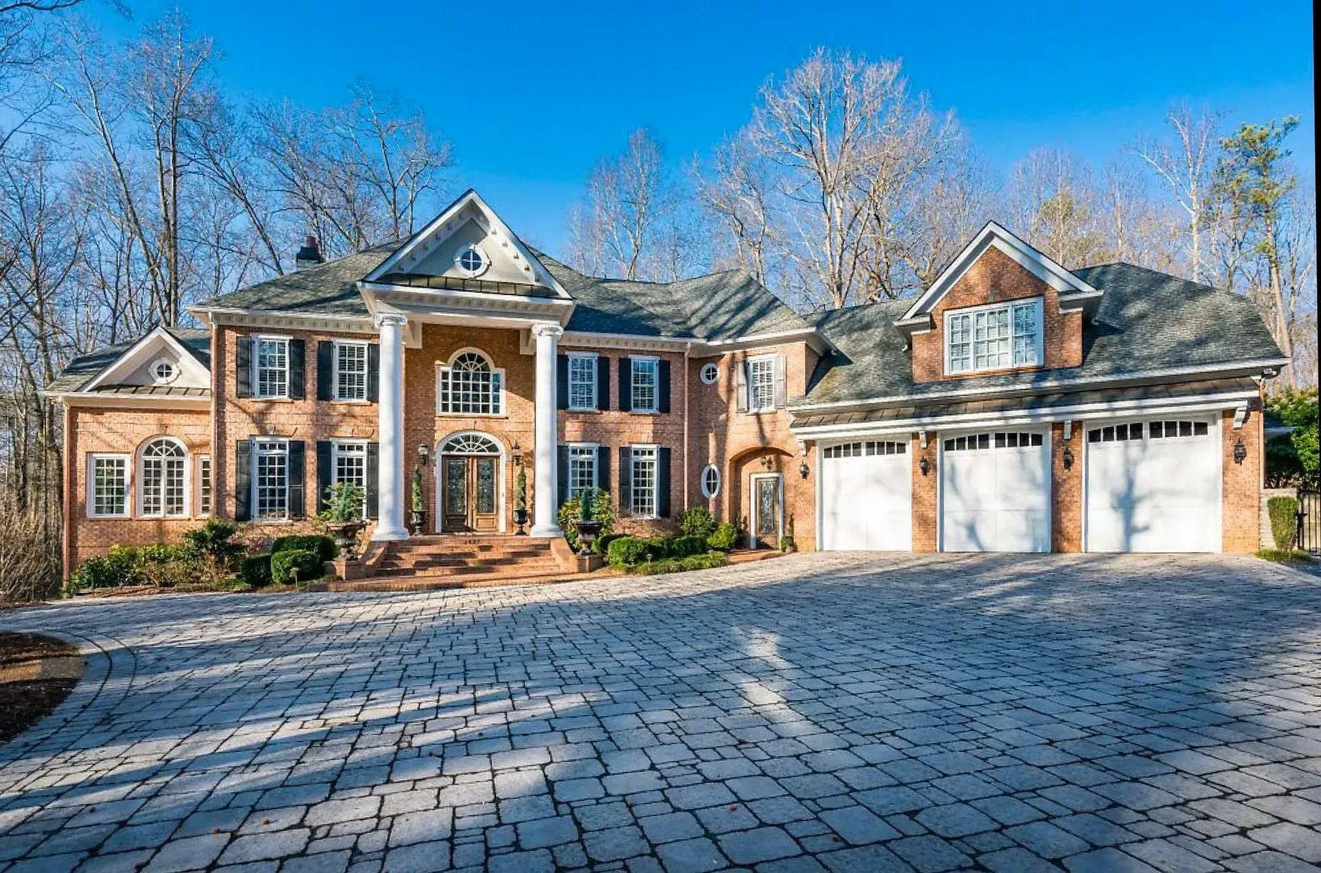 Large brick suburban house with three-car garage, stone driveway, and leafless trees in front yard