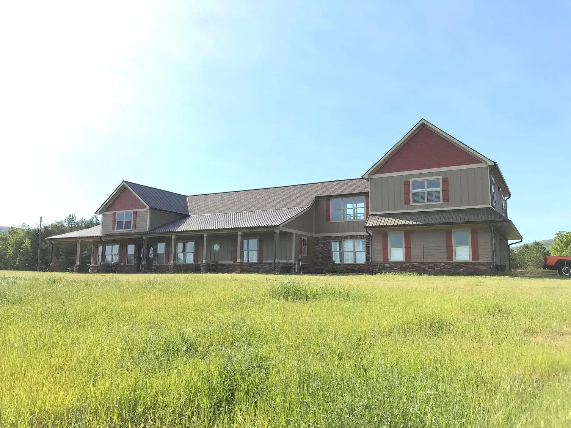 Wide farmhouse with brown siding and red trim in a grassy field under a bright blue sky.