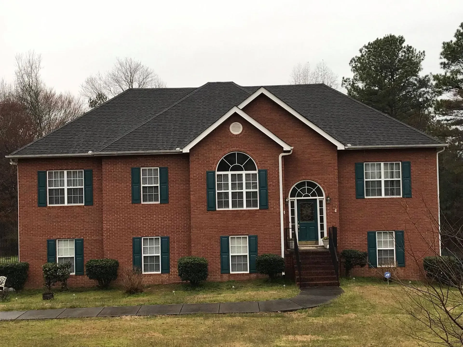 Two-story red brick house with green shutters and a black roof, front steps, and a small lawn
