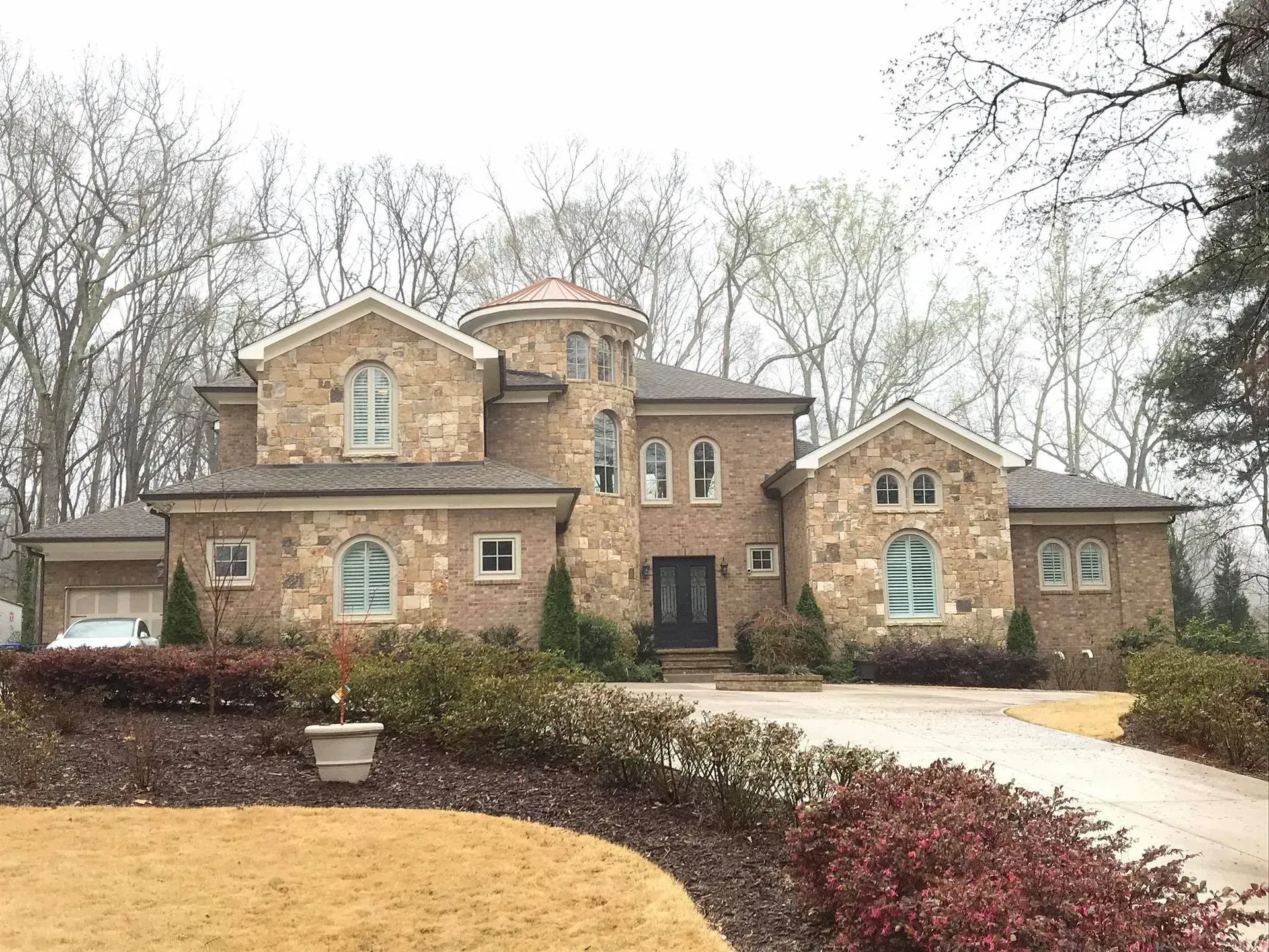 Large stone suburban house with a manicured front yard and a curved driveway under overcast skies