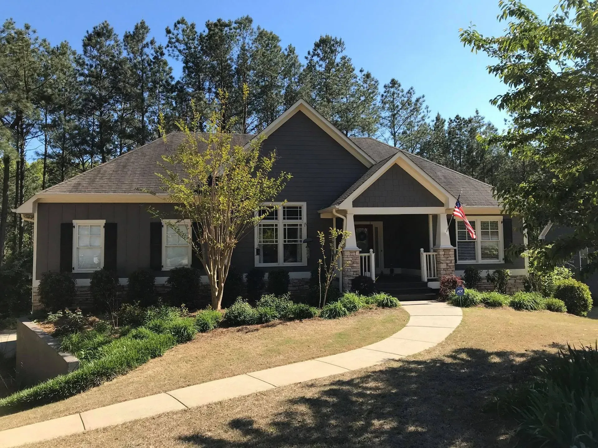 Single-story dark gray house with a curved front walkway and an American flag, surrounded by trees and shrubs