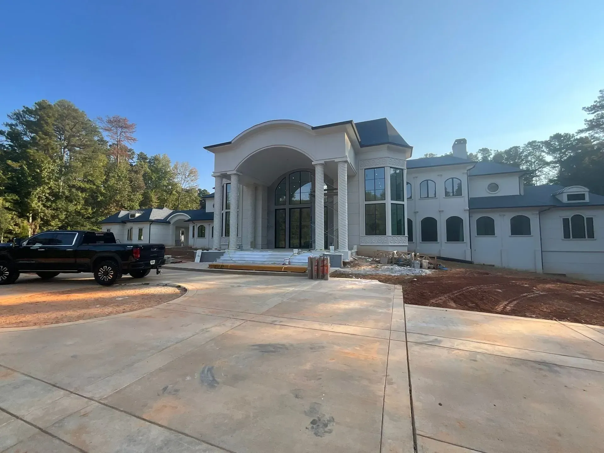Large unfinished white mansion with columns and a wide concrete driveway under a clear blue sky