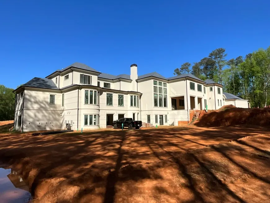Large white multi-story house under construction on a cleared lot with red dirt and trees under a blue sky