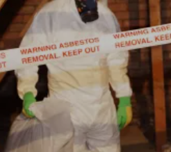 Person in white hazmat suit and green gloves behind asbestos warning tape, in a brick doorway.