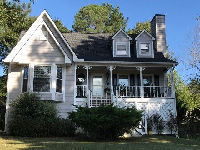 White two-story house with a front porch, dormer windows, and a brick chimney surrounded by trees
