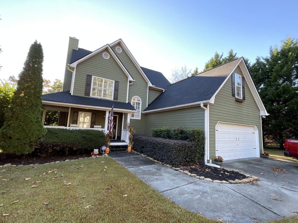 Suburban two-story house with green siding, white garage, front porch, and driveway on a sunny day