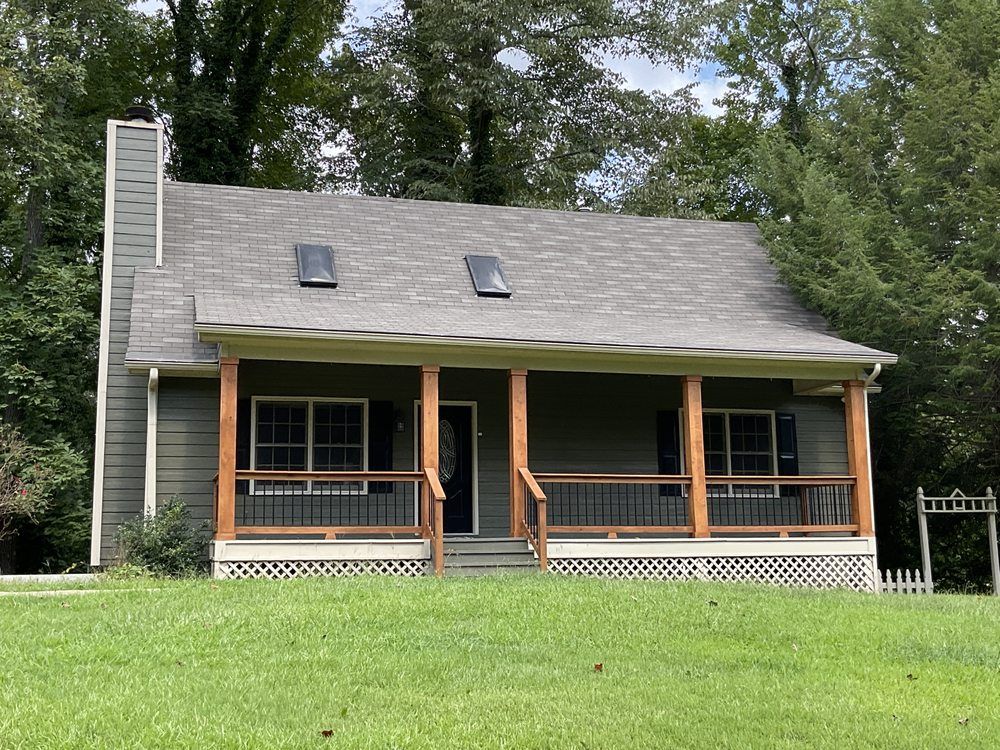 Small gray house with a front porch, stone chimney, and green lawn surrounded by trees.