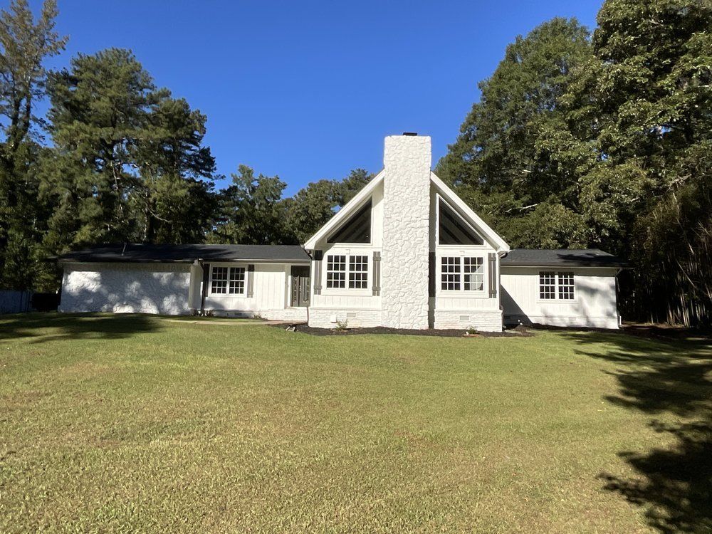 White house with a tall central chimney, black shutters, and a large green lawn under a blue sky.