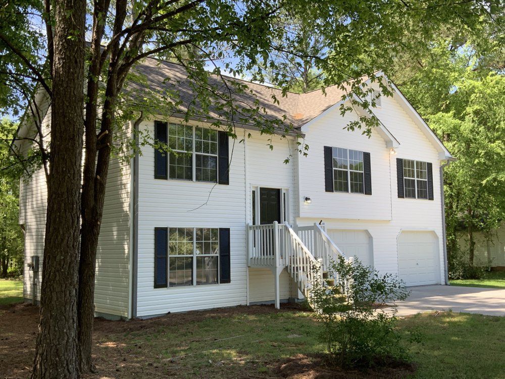 White two-story house with black shutters, front porch, and attached garage among trees