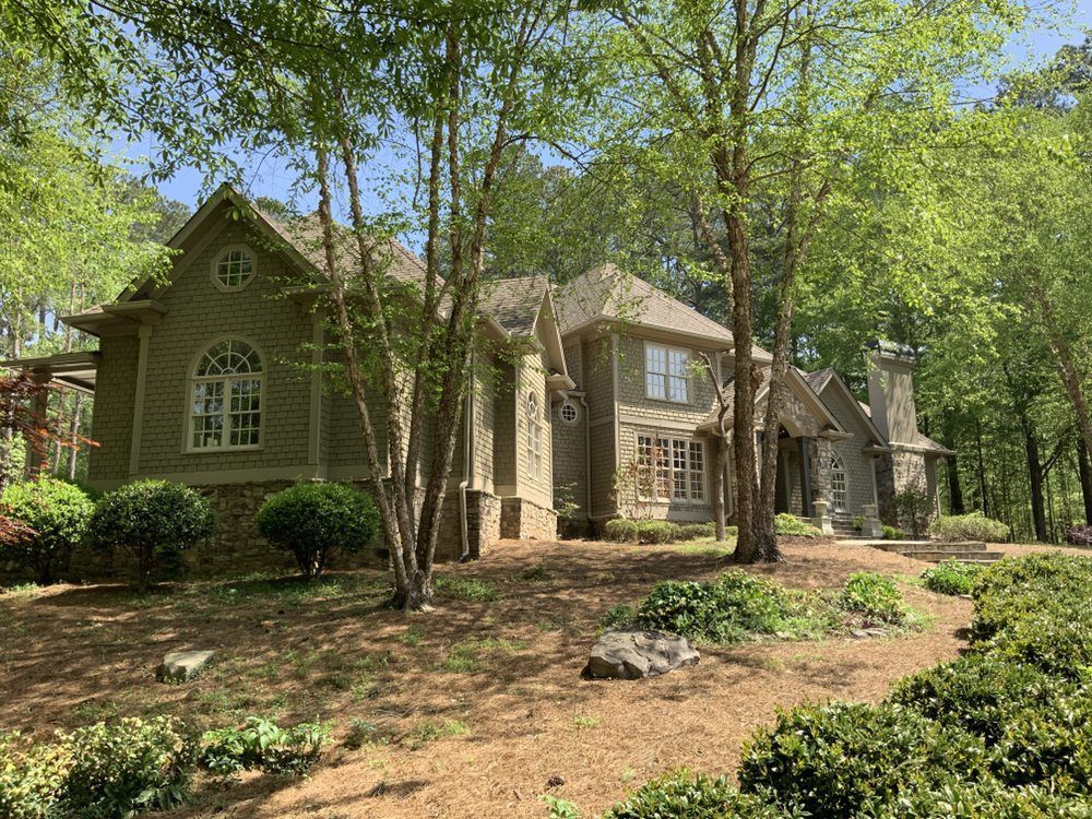 Large stone and wood house in a leafy wooded yard with trees and shrubs.