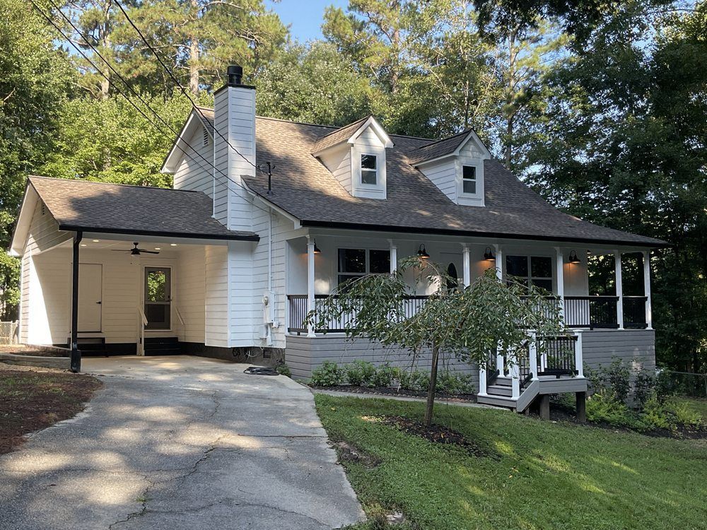 White house with porch and dormer windows, front driveway, surrounded by trees and lawn