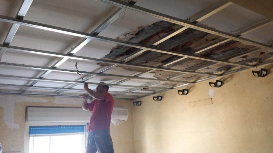 Uomo che installa un controsoffitto. Il soffitto è parzialmente rimosso, rivelando travi in legno. Pareti gialle.